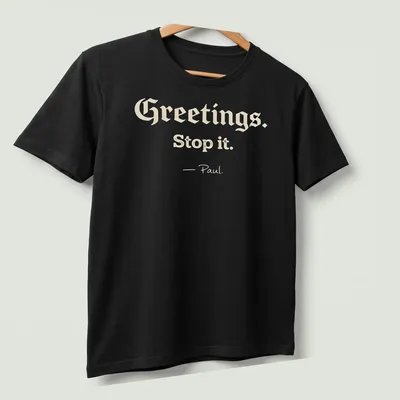 Playful script with Christian humor saying 'Greetings. Stop it. - Paul.' on a cream background — printed on a unisex t-shirt (angled view)