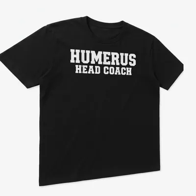 White T-shirt with bold text 'HUMERUS HEAD COACH' and anatomical humerus bone graphic — printed on a unisex t-shirt (angled view)